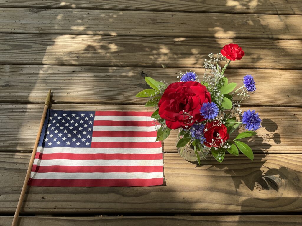 A bouquet with a red rose, white baby's breath, and blue bachelor buttons next to an American Flag